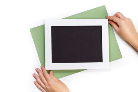 hands holding horizontal white photo frame with black field and green paper under angle on white background isolated with real shadowsの写真素材