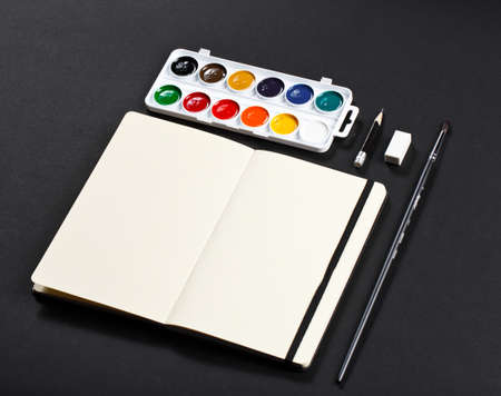 Open blank notepad with empty white pages laying with water-color palette brush pencil eraser on black background. ideal for textの写真素材