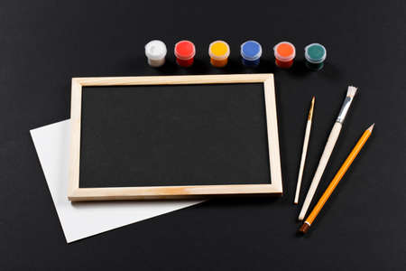 wooden frame with black field and with brushes and set of gouache of different colors on black paper. Mockup. Real photoの写真素材