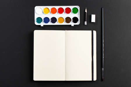 Open blank notepad with empty white pages laying with water-color palette brush pencil eraser on black background. ideal for textの写真素材
