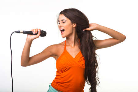 The brunette with long hair is singing on the white background. Pretty lady in orange shirt with microphone.の写真素材