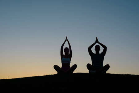 Morning yoga on the nature. Silhouette figures of people at sunset. People relax. Rest at nature. Boy and girl meditating at sunset.の写真素材