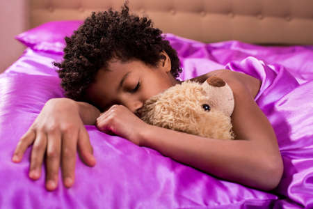 Afro boy sleeping with teddybear. Kid in bed with teddybear. Peaceful atmosphere of home. There's never too much comfort.の写真素材