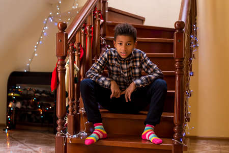 Mulatto boy in colorful socks. Young boy sits on staircase. When holidays come near. Few hours before celebration.の写真素材