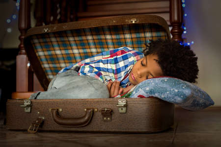 Afroamerican kid sleeps inside suitcase. Young boy's nap inside suitcase. He needs some rest. Holidays make him tired.の写真素材