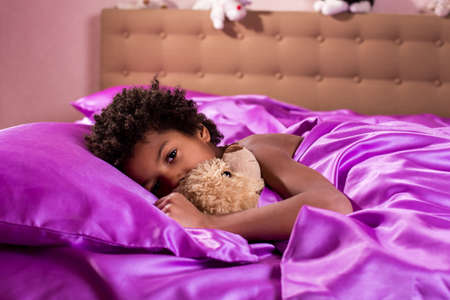 Afro boy with teddy bear. Boy in bed with teddybear. Tired look of his eyes. It's time to slip.の写真素材