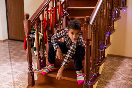 Black kid sitting on staircase. Boy at home during Christmas. Waiting for guests to come. Feeling bored on holiday.の写真素材