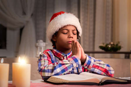 Darkskinned boy praying on Christmas. Little Santa prays to God. Purity of young soul. Prayer after reading Bible.の写真素材