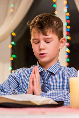 Boy praying at the table. Kid prays at nighttime. Christmas prayer at city church. Youthful...