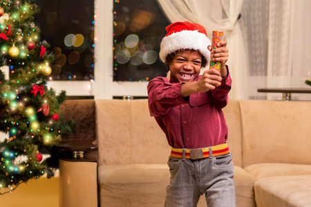 Afro boy holding Christmas petard. Little Santa with fire cracker. It will be loud. Happy holidays, dear neighbours.の写真素材