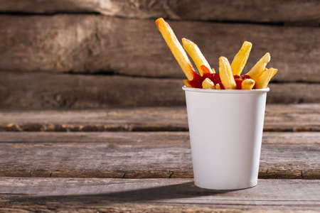 Fries with sauce in cup. French fries with tomato ketchup. Aged table with tasty snack. Popular fast...
