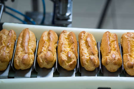 Conveyor line with eclairs. Baked pastry laying on conveyor. Dessert with cream filling. Crispy food...