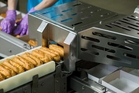Eclair shells and white boxes. Eclairs on conveyor beside worker. Complex machine at confectionery...