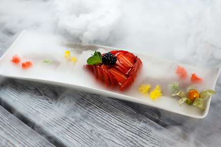 Mint leaf on sliced strawberries. Dessert on long white plate. Flan served with dry ice. High-calorie dish with fruits.の写真素材