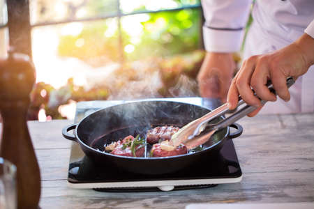 Hand with tongs touches meat. Steam coming from frying pan. Chef roasts beef. Smells very appetizing.の写真素材
