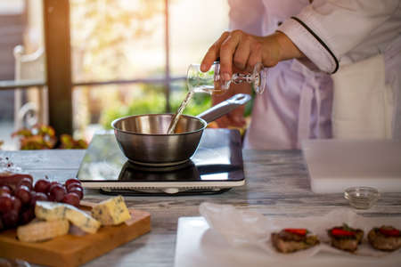 Glass pouring liquid on saucepan. Male hand near small cooker. Chef prepares roquefort sauce. Add a bit of wine.の写真素材