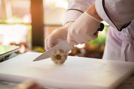 Hand with knife cuts garlic. Vegetable on white cooking board. Need a special ingredient. Chef knows his work well.の写真素材