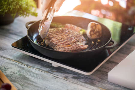 Tongs touch brown meat. Black round pan. Roasted beef for lunch. Portable cooker in the kitchen.の写真素材