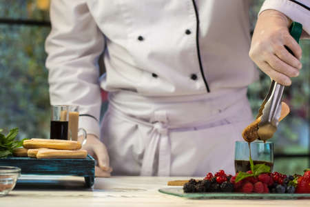 Hand with tongs holding biscuit. Glass with dark liquid. Chef dips savoiardi into coffee. Mastery of...
