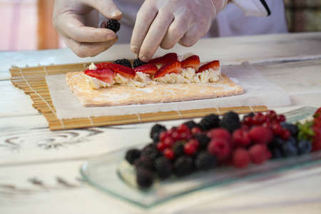 Hand in glove holds berry. Dough and cream cheese. Chef is making sweet rolls. Best recipe of summer dessert.の写真素材