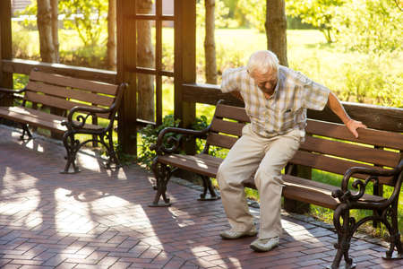 Senior man holding his back. Old male near park bench. Strong pains in spine. Need of medication and therapy.の写真素材