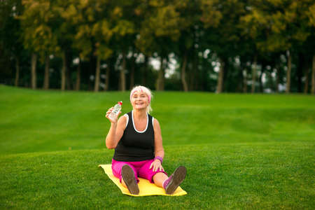 Senior lady sitting on mat. Woman smiling and holding bottle. The joy of being healthy. Take a sip.の写真素材