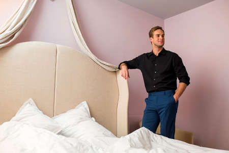 Young man standing near bed. Smiling guy in black shirt. Be elegant and enchant women.の写真素材