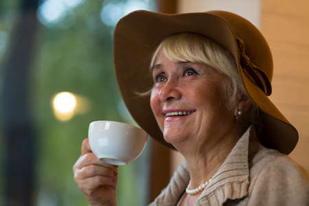 Senior woman holding white cup. Elderly lady in hat smiling. Few sips of cappuccino.の写真素材