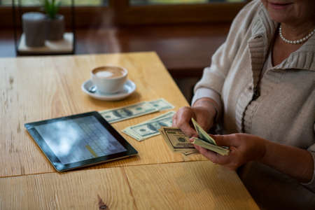 Senior woman holding dollars. Money and tablet on table. Income of private business.の写真素材