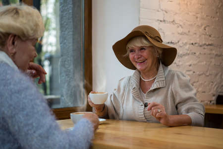 Senior woman with coffee smiling. Two ladies near cafe window. Morning conversation with a friend.の写真素材