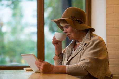 Elderly woman with tablet. Senior lady holding coffee cup. Create account in social network.の写真素材