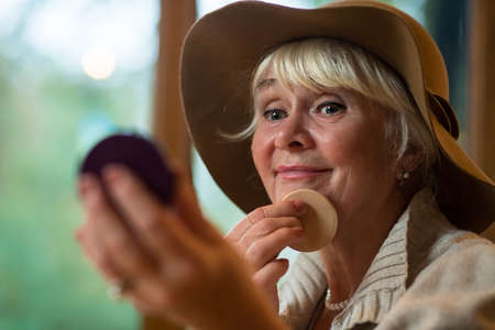 Elderly woman applying makeup. Senior lady in a hat. Strive to perfection.の写真素材