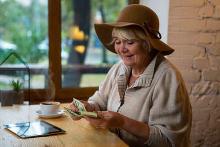 Senior woman holding money. Coffee and tablet on table. Count all savings.の写真素材