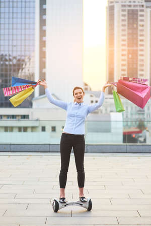 Happy woman with colorful bags. Female on gyroboard, city. Successful shopping day.