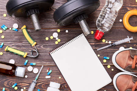 Empty notebook, syringes, pills, dumbbells. Pharmaceutical preparations and sport equipment on wooden background.の写真素材