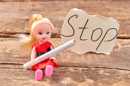 Toy doll holds a cigarette. Doll with cigarette, message stop, old wooden background. Metaphor concept stop smoking.の写真素材