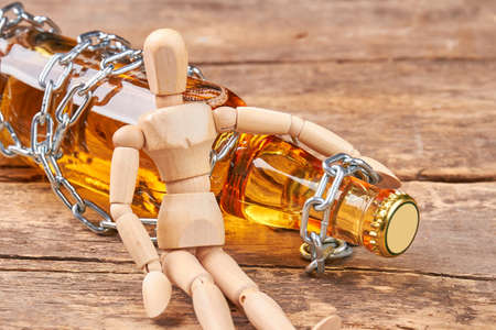 Human wooden dummy sitting with bottle. Bottle wrapped in chain, wooden human mannequin, old wooden background. Concept of alcohol addiction.の写真素材