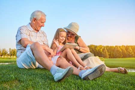 Smiling girl with grandparents, tablet. Happy people sitting on grass. How internet makes life easier.の写真素材