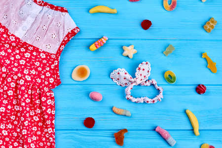 Summer kids wear, sweets and head wrap. Red small girls dress with pattern of white flowers, hairband, sweets, blue wooden background.の写真素材