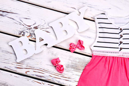Text baby, baby dress, hair accessories. Cute brand dress for baby-girl, pink hair bows, old wooden background. Infant girl summer fashion background.の写真素材