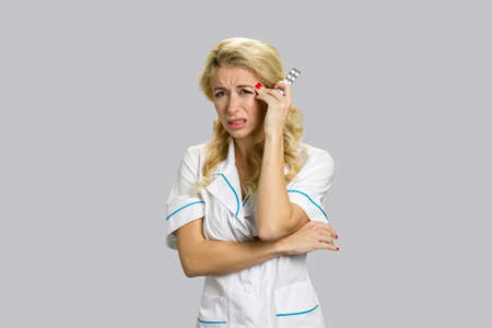 Upset nurse suffering from headache. Overworked young female doctor having strong headache and standing with pills on grey background.の写真素材