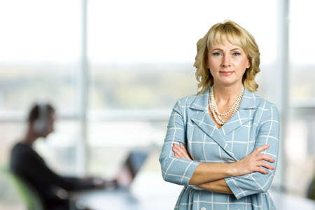 Mature woman on office window background. Beautiful business lady with crossed arms stading on blurred background.の写真素材