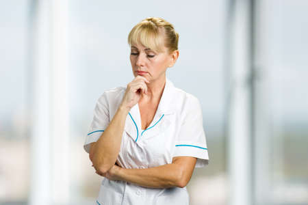 Confident thinking mature nurse. Portrait of thoughtful mature medical doctor holding chin on hand and thinking on blurred background.の写真素材
