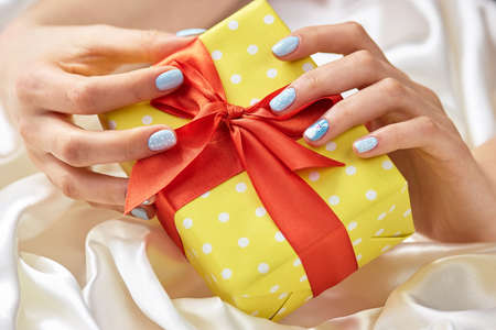 Gift box in female manicured hands. Beautiful yellow gift box in woman hands with gentle winter manicure. Holidays and celebration concept.の写真素材