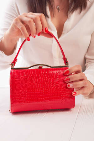 Female hands holding red handbbag. Woman with beautiful manicure holding bright lacquered clutch on white wooden table.の写真素材
