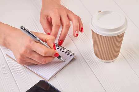 Woman manicured hands writing in notebook. Gentle female hand with beautiful manicure writing with pen in empty notebook. Writing woman hand, paper notebook, disposable coffee cup.の写真素材