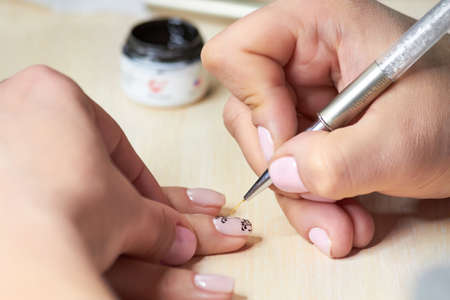 The drawing process on female nails. Female manicurist hand putting black lace on nude nail polish in beauty salon. Female nails manicure.の写真素材