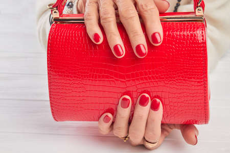 Elegant red handbag in female hands. Elderly woman hands with red manicure holding lacquered handbag close up. Womans modern accessory.の写真素材
