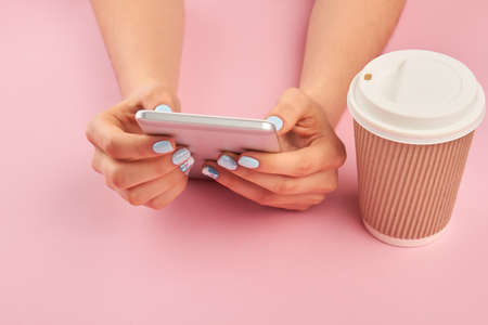 Smartphone in female manicured hands. Hands with manicure holding mobile phone, cardboard cup of coffee, pink background.の写真素材