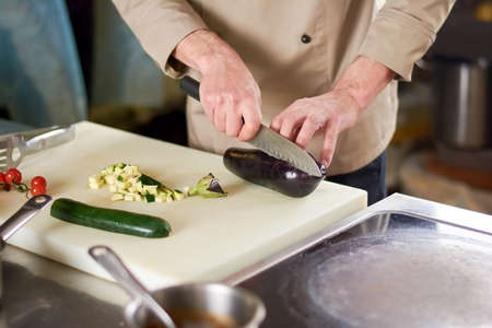 Chef cutting eggplant by steel knife in restaurant. Male chef cutting eggplant on cutting board next to sliced zucchini. European cuisine concept.の写真素材